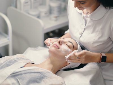 Woman lying during cosmetic procedure while skillful female cosmetologist making facial treatment